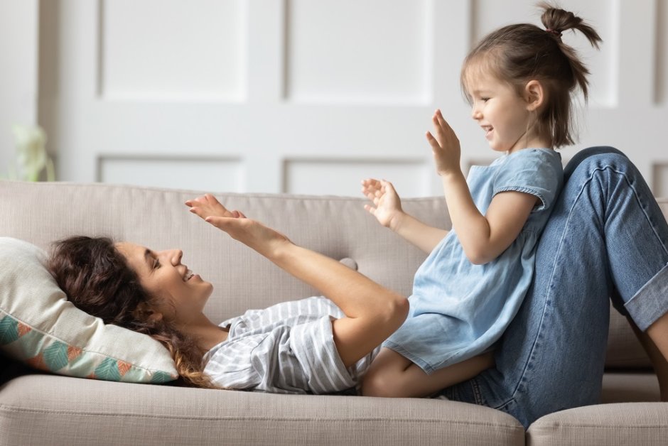 Smiling mother playing with little daughter, clapping hands on couch